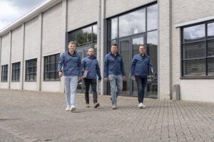 Vier mannen in blauwe bedrijfskleding lopen samen naar buiten vanaf een modern industrieel gebouw met grote ramen. Twee van hen dragen laptops. Het is een bewolkte dag.